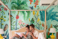 Couple relaxing on a white canopy bed with breakfast in bed, set against colorful tropical wallpaper at Boardwalk Boutique Hotel Aruba.