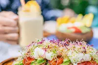 Avocado toast from Coco Cafe, topped with shredded vegetables on a wooden board, with a smoothie and a blurred guest at an outdoor table.