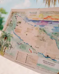 Hands holding an illustrated treasure map on the beach at Boardwalk Boutique Hotel Aruba, with sea and palm trees in the background.