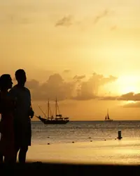Couple silhouette watching the sunset on Palm Beach, Aruba, with calm sea and sailboats in the distance.