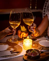 Two guests clink glasses of white wine at a candlelit table at The Coco Cafe, Boardwalk Boutique Hotel Aruba, with small dishes on the table.
